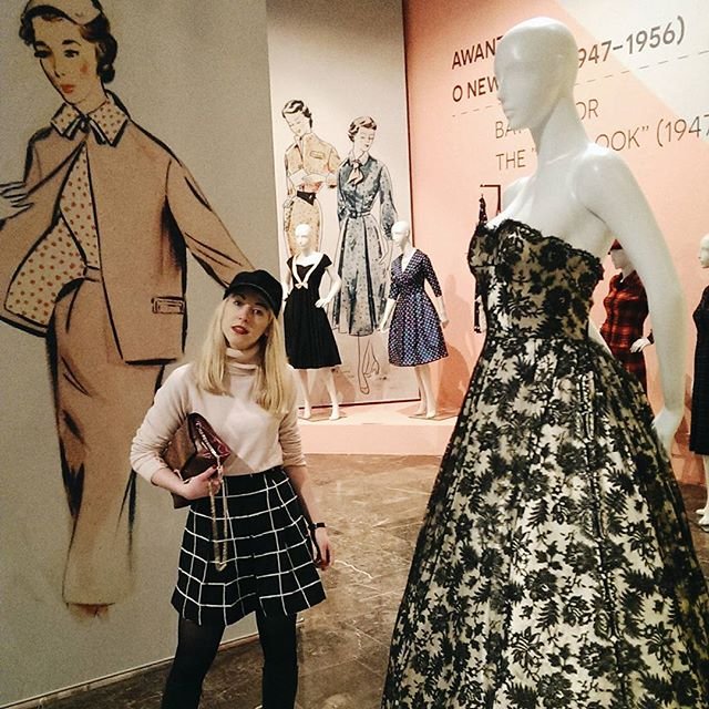 Kobieta w czarnej czapce i jasnym swetrze trzymająca torebkę stoi na wystawie mody obok dużej manekiny w długiej czarno-białej sukni z kwiatowym wzorem, a w tle widać ilustracje modowe i kilka mniejszych manekinów na różowo-beżowej ścianie.