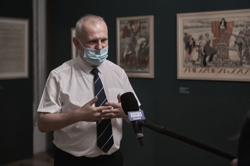 Mężczyzna w białej krótkorękawowej koszuli z granatowym krawatem i jednorazową maseczką na twarzy, gestykulujący przed mikrofonem oznaczonym TVP3, stoi w sali wystawowej z oprawionymi obrazami na ciemnej ścianie.
