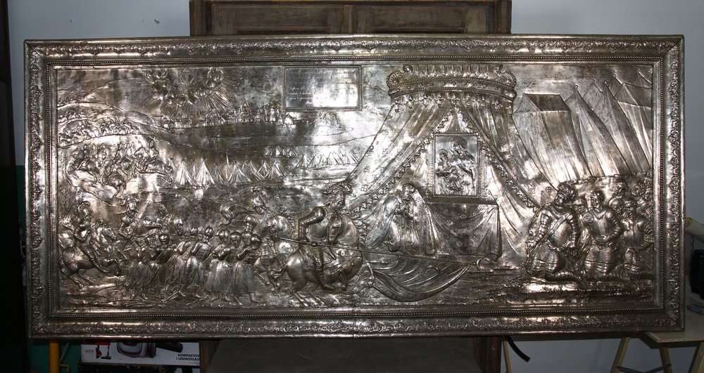 Poziomy lśniący srebrny relief w ozdobnej ramie przedstawiający gęsto wypełnioną scenę z ugrupowaniami uzbrojonych postaci i konną kawalerią po lewej, w tle rzędy namiotów i łodzie z żaglami po prawej oraz centralnie duży zdobiony baldachim z zasłonami skrywający postać, wszystko wykonane w technice wybicia metalu z bogatymi detalami.