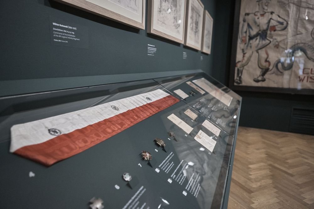 Witryna wystawowa w muzeum z oszklonym blatem, na którym leży długi biało-czerwony pas materiału oraz rozłożone odznaki, dokumenty i opisy, nad nią oprawione rysunki na ciemnej ścianie i po prawej duże malowane płótno, parkietowa podłoga.