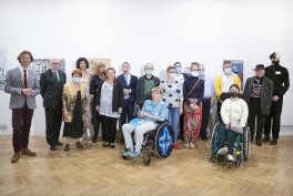 Kilkanaście osób ustawionych obok siebie w sali wystawowej z białymi ścianami i wiszącymi obrazami, część stoi, dwie osoby siedzą na wózkach inwalidzkich, kilka osób ma maseczki, na podłodze widoczny drewniany parkiet.
