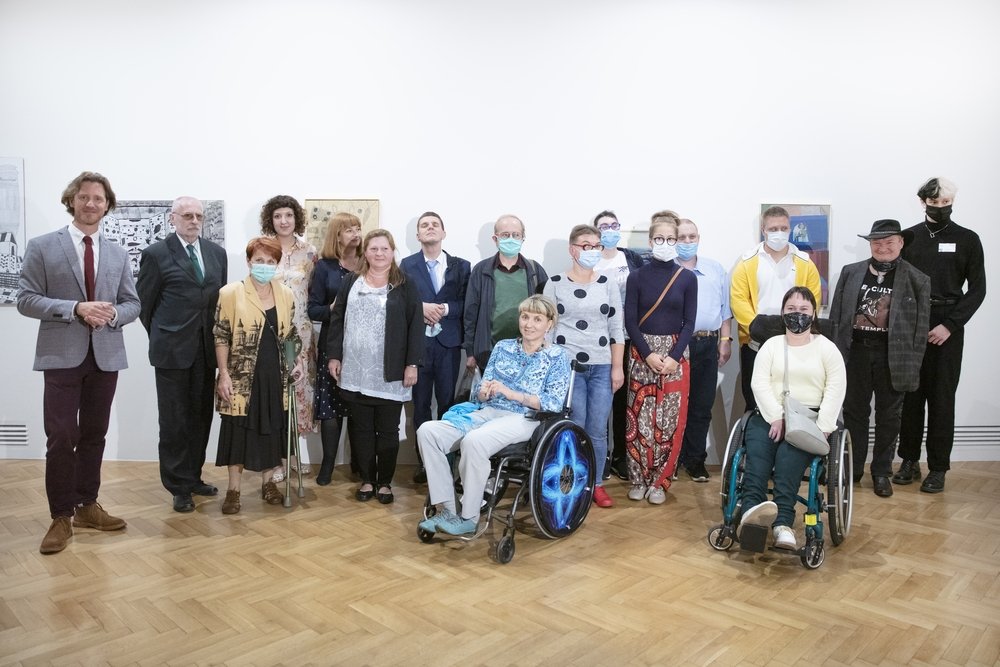 Grupowe zdjęcie kilkunastu osób — mężczyzn i kobiet — ustawionych w jednym rzędzie w jasnej sali wystawowej z białymi ścianami i kilkoma obrazami, na drewnianej parkietowej podłodze; wśród nich dwie osoby na wózkach inwalidzkich, kilka osób w maseczkach ochronnych i jedna osoba z laską.