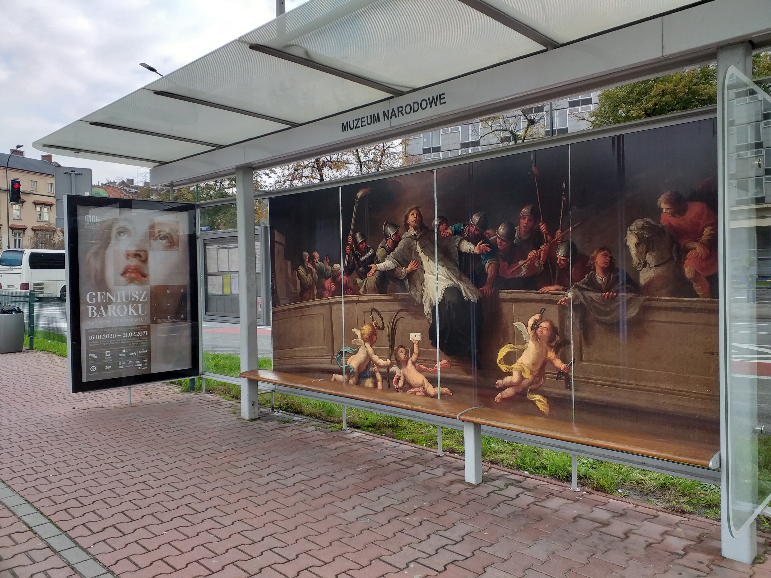 Wiata przystankowa oznaczona napisem MUZEUM NARODOWE z przeszklonym dachem i drewnianą ławką, na tylnej szybie wielkoformatowa reprodukcja barokowego obrazu z centralną postacią w białej szacie otoczoną żołnierzami, kilkoma puttami w pierwszym planie i koniem po prawej, a po lewej wewnętrzny panel z plakatem wystawy "GENIUSZ BAROKU" oraz chodnik z czerwonej kostki i budynki w tle.