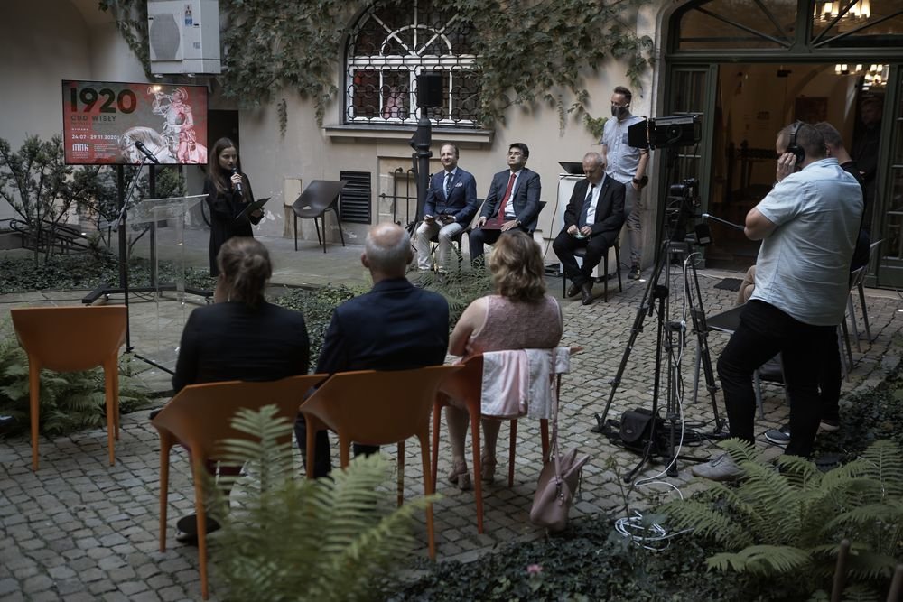 Kobieta trzymająca mikrofon przy przezroczystym pulpicie przemawiająca na małym dziedzińcu przed kilkoma siedzącymi słuchaczami, za nią rząd czterech mężczyzn w garniturach, po prawej kamera na statywie z operatorem i otwarte drzwi do budynku, po lewej ekran z napisem "1920", a całość na brukowanej nawierzchni z paprociami i porośniętymi bluszczem ścianami.