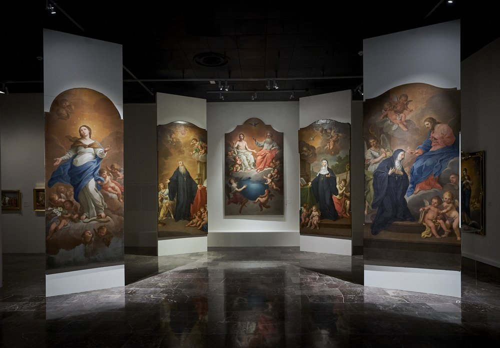 Ciemna galeria z połyskującą marmurową podłogą, w której w półokręgu ustawiono wysokie wolnostojące panele z dużymi obrazami religijnymi przedstawiającymi postacie na chmurach oraz anioły i cherubinki, z centralnym panelem z siedzącymi postaciami.