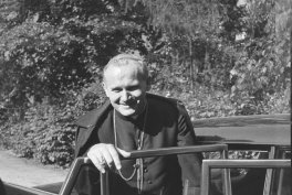 Czarno-białe zdjęcie mężczyzny o siwych włosach w sutannie z krzyżem na piersi, uśmiechającego się i opierającego się na otwartym drzwiach samochodu, z drzewami w tle.