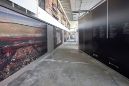 Wnętrze przestrzeni wystawowej z długim korytarzem; po lewej nisko zawiesione panoramiczne fotografie, po prawej wysokie czarne panele z białymi napisami, betonowa posadzka i wysoki sufit z metalowymi belkami i oświetleniem.