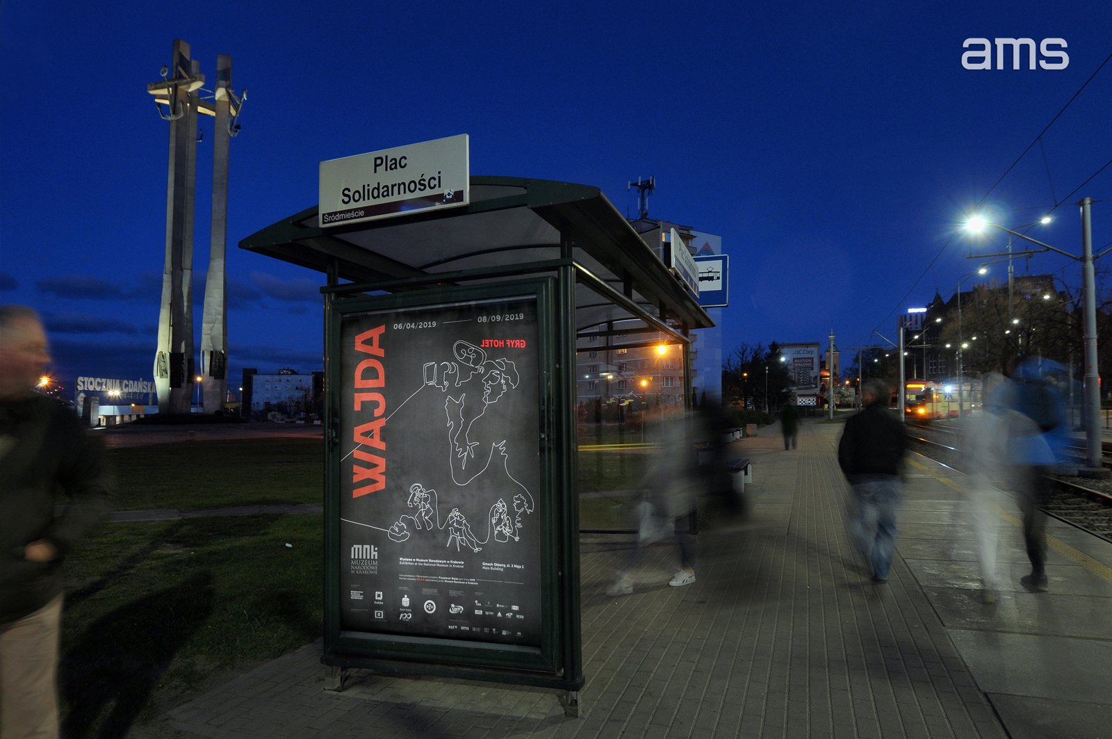 Nocna scena placu z przystankiem tramwajowym i zadaszoną wiatą z dużym czarnym plakatem z czerwonym napisem "WAJDA", nad wiatą tablica z napisem "Plac Solidarności", po lewej dwie wysokie metalowe kolumny, rozmyci piesi idący po chodniku, tory i nadjeżdżający tramwaj z prawej oraz oświetlone ulice i logo "ams" w prawym górnym rogu.