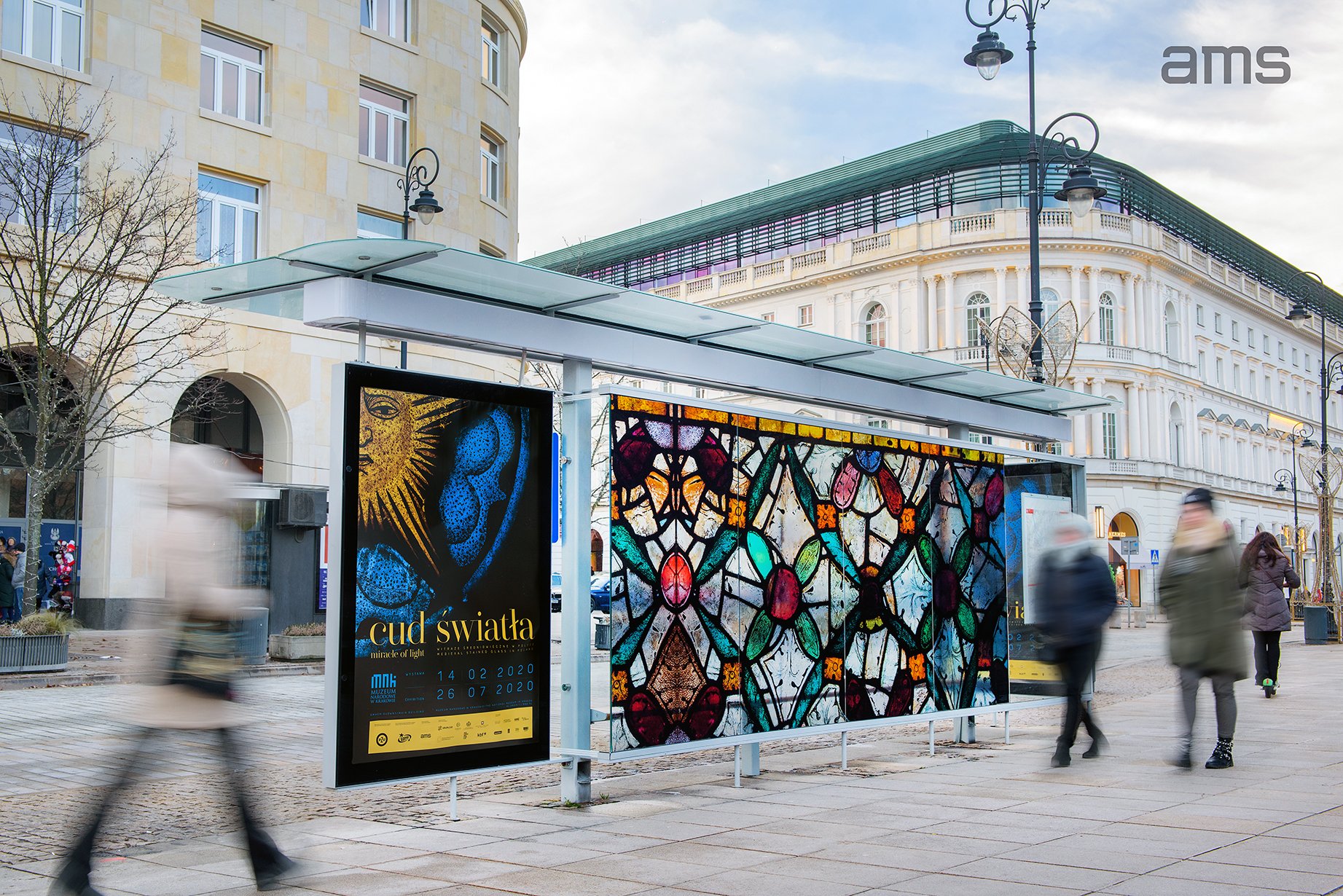 Wiata przystankowa z przeszklonym dachem i trzema panelami reklamowymi — lewy panel z plakatem „cud swiata” z motywem słonecznym i datami, środkowy panel z kolorowym wzorem przypominającym witraż, rozmyci przechodnie na chodniku, w tle jasny klasycystyczny budynek z zielonym dachem, latarnie uliczne, bezlistne drzewa i logo „ams” w prawym górnym rogu.