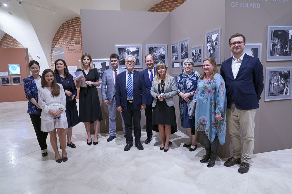 Grupa osób w różnym, częściowo formalnym ubiorze stoi w rzędzie w przestronnej sali wystawowej z łukowym stropem i odsłoniętą czerwoną cegłą, na ścianach za nimi w ramkach wyeksponowane czarno-białe zdjęcia, a pod nimi jasna, polerowana podłoga.