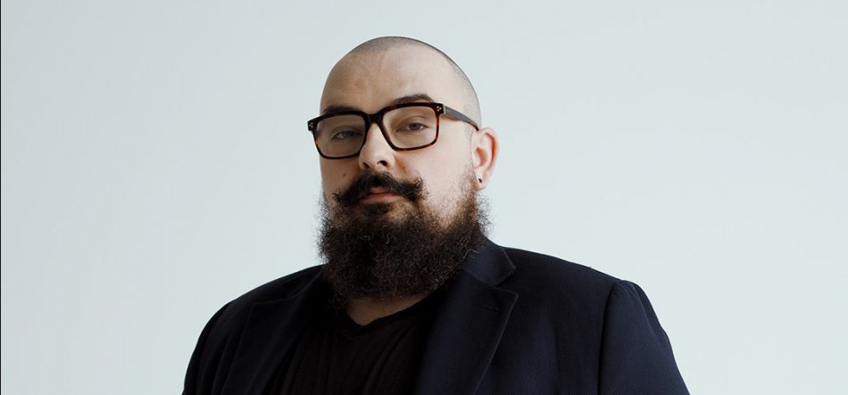 Półpostać mężczyzny o ogolonej głowie, gęstej brodzie i wąsach, w dużych ciemnobrązowych okularach i z kolczykiem w lewym uchu, ubrana w czarną koszulkę i ciemną marynarkę na jasnoszarym tle.