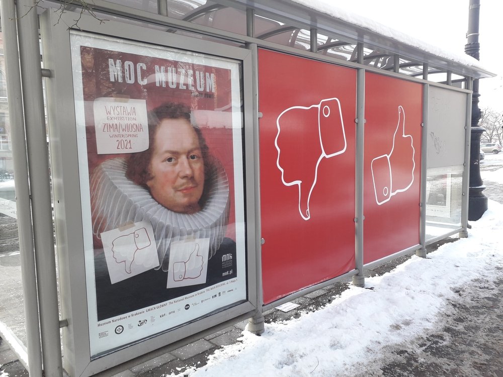 Przystanek autobusowy z przeszklonym panelem reklamowym: po lewej plakat z portretem osoby w kryzie i napisem „MOC MUZEUM” oraz datą „ZIMA/WIOSNA 2021”, po prawej czerwone panele z białymi konturami kciuka w dół i w górę, wokół widoczny śnieg.