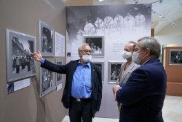 Trzech mężczyzn w marynarkach i jednorazowych maseczkach stoi w galerii przed ścianą z oprawionymi czarno‑białymi fotografiami, a jeden z nich wskazuje ręką na jedną z fotografii; w tle większa czarno‑biała fotografia i sufitowe oświetlenie wystawy.