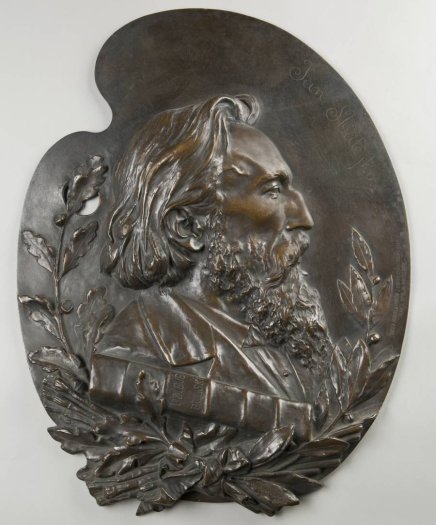 Brązowy relief w kształcie palety malarskiej przedstawiający prawy profil starszego mężczyzny z długimi włosami i bujną brodą, otoczony gałązkami laurowymi i dębowymi oraz z widoczną książką przy piersi.