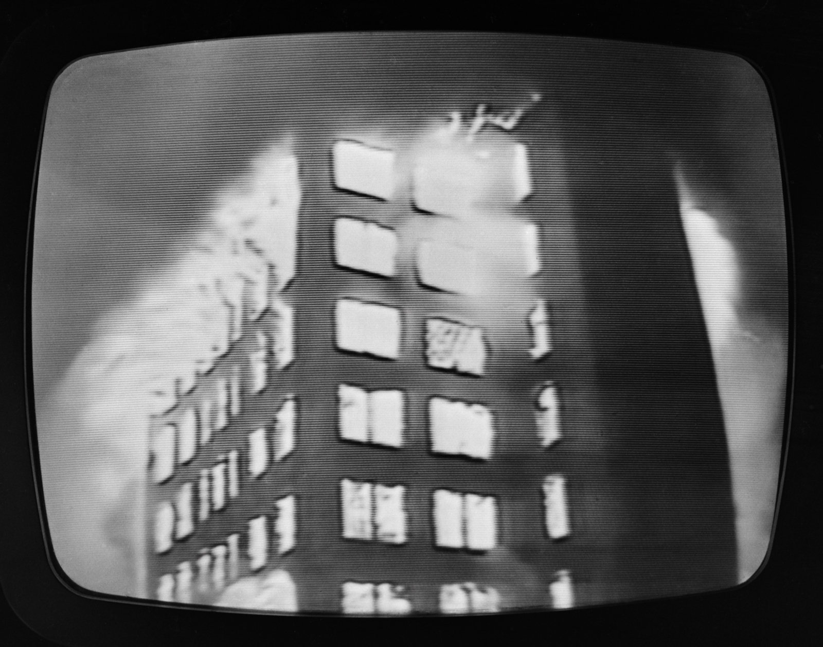 Czarno-biały, ziarniasty obraz na ekranie telewizora CRT przedstawiający ukośnie ustawiony wielopiętrowy budynek z regularnymi kwadratowymi oknami, jasnymi, rozmytymi plamami i smugami przy górnych kondygnacjach sugerującymi dym lub refleksy świetlne oraz widoczne poziome linie skanowania i czarne obramowanie kineskopu.