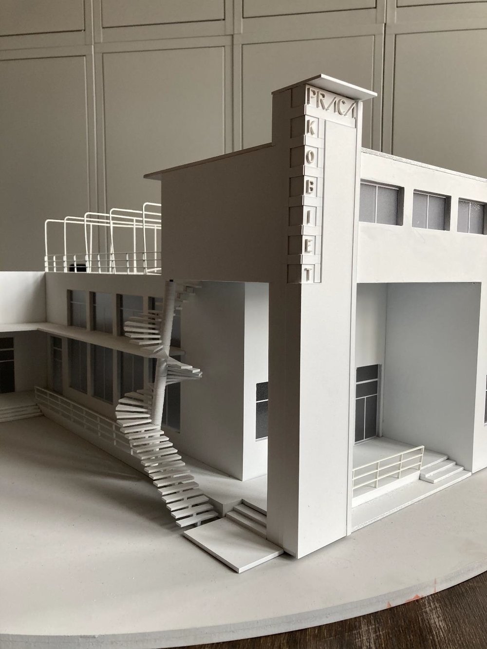Biały model architektoniczny budynku z zewnętrzną spiralną klatką schodową, tarasem z balustradą, prostokątnymi oknami, wejściowymi schodami i pionowym szyldem na narożniku z literami PRACA i KOBIET.