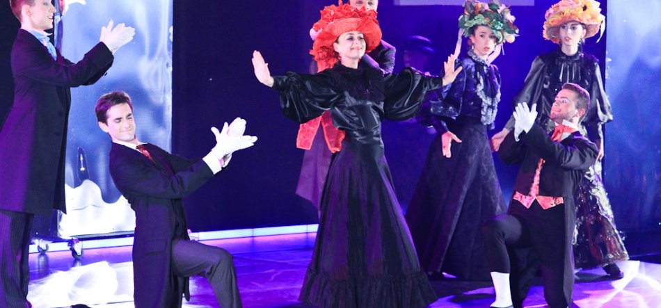 Grupa aktorów na scenie w stylizowanych czarnych strojach — dwaj mężczyźni w frakach i białych rękawiczkach klęczą po bokach, kilka kobiet w długich ciemnych sukniach stoi w tle w dużych dekoracyjnych kapeluszach (czerwonym, zielonym i żółtym), centralna kobieta w czerwonym kapeluszu ma uniesione ramiona, scena oświetlona fioletowo‑niebieskim światłem.