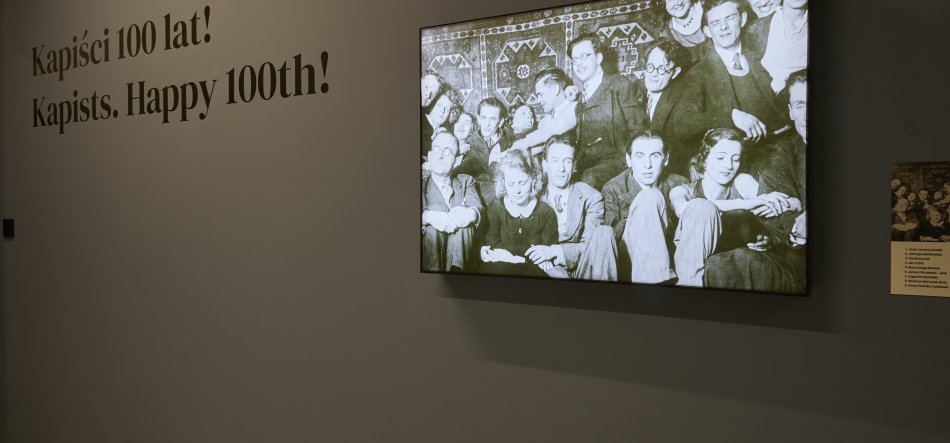 Szara ściana wystawowa z napisem Kapiści 100 lat! Kapists. Happy 100th! po lewej oraz podświetlonym czarno‑białym zdjęciem grupowym w ramie i małą tabliczką informacyjną po prawej.