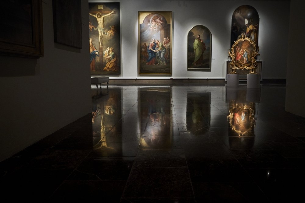 Wnętrze ciemnej galerii z kilkoma oświetlonymi obrazami o tematyce religijnej — po lewej duże przedstawienie Ukrzyżowania, centralnie duże płótno ze sceną grupową, obok wąskie łukowate przedstawienie i po prawej ozdobna złocona rama z obrazem, a wszystkie prace odbijają się na błyszczącej marmurowej podłodze.
