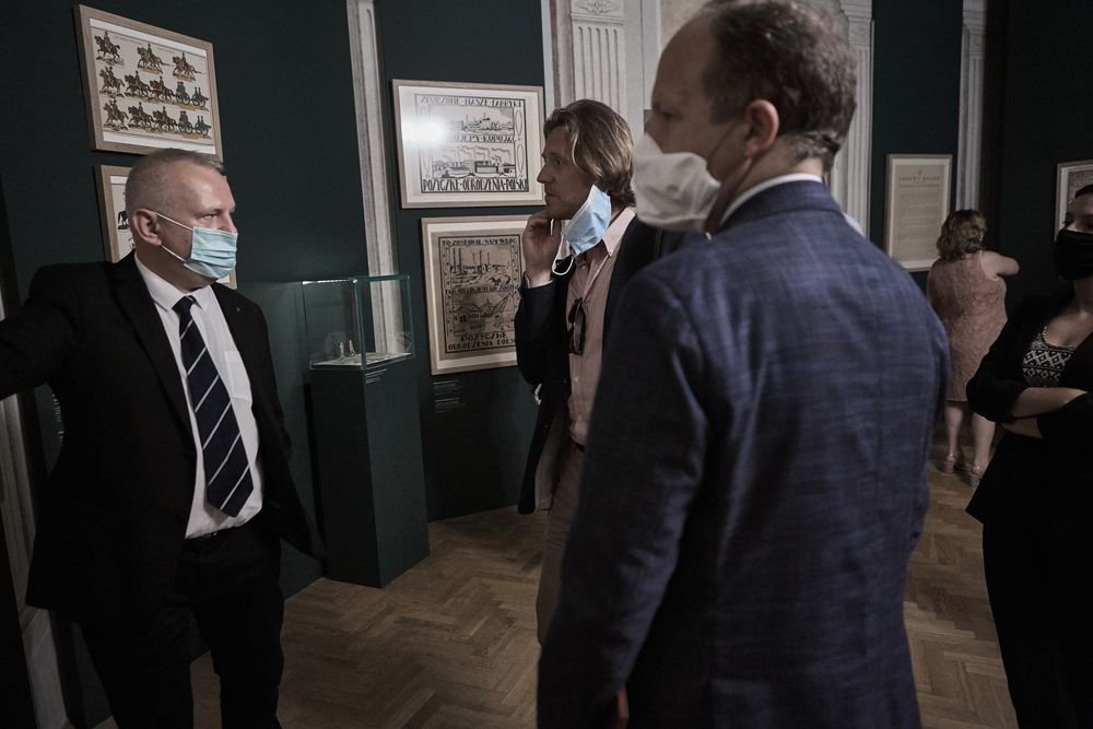 Trzech mężczyzn w maskach stoi blisko siebie w sali wystawowej z ciemnozielonymi ścianami, oprawionymi grafikami na ścianach, oszkloną gablotą i drewnianą podłogą, a w tle widać jeszcze dwie osoby przy ścianie.