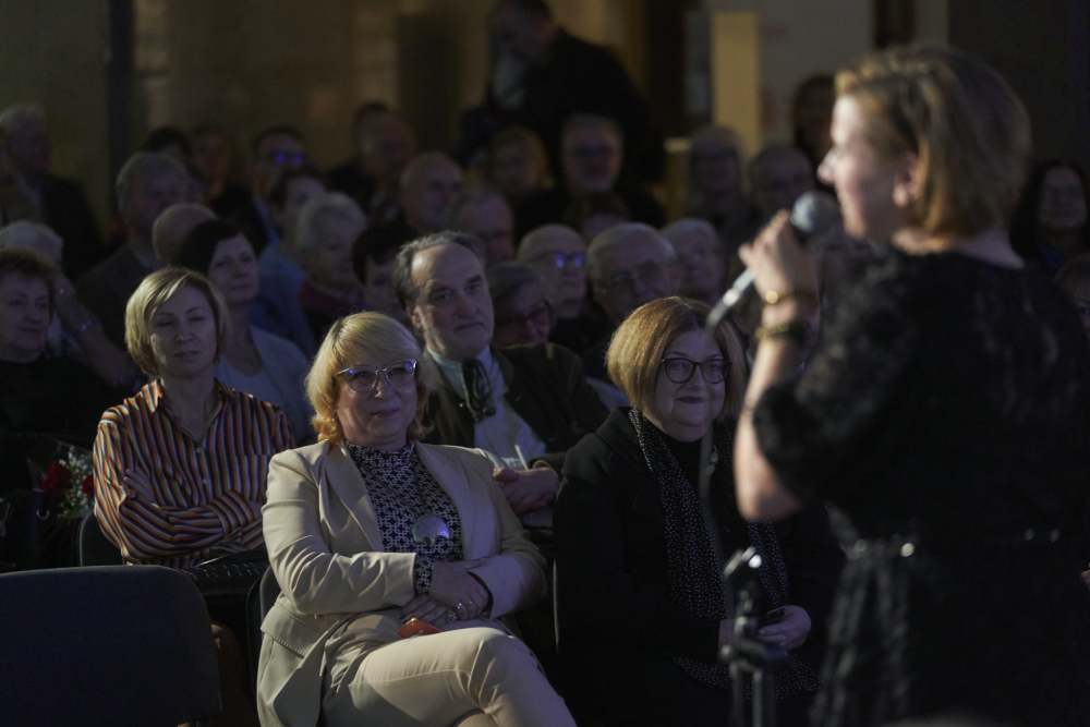 Widok publiczności w sali — w ostrości dwie kobiety na pierwszym rzędzie siedzące obok siebie (jedna w jasnym garniturze i okularach, druga w ciemnym płaszczu i okularach), za nimi kilka rzędów starszych osób, po prawej na pierwszym planie rozmyta osoba trzymająca mikrofon.