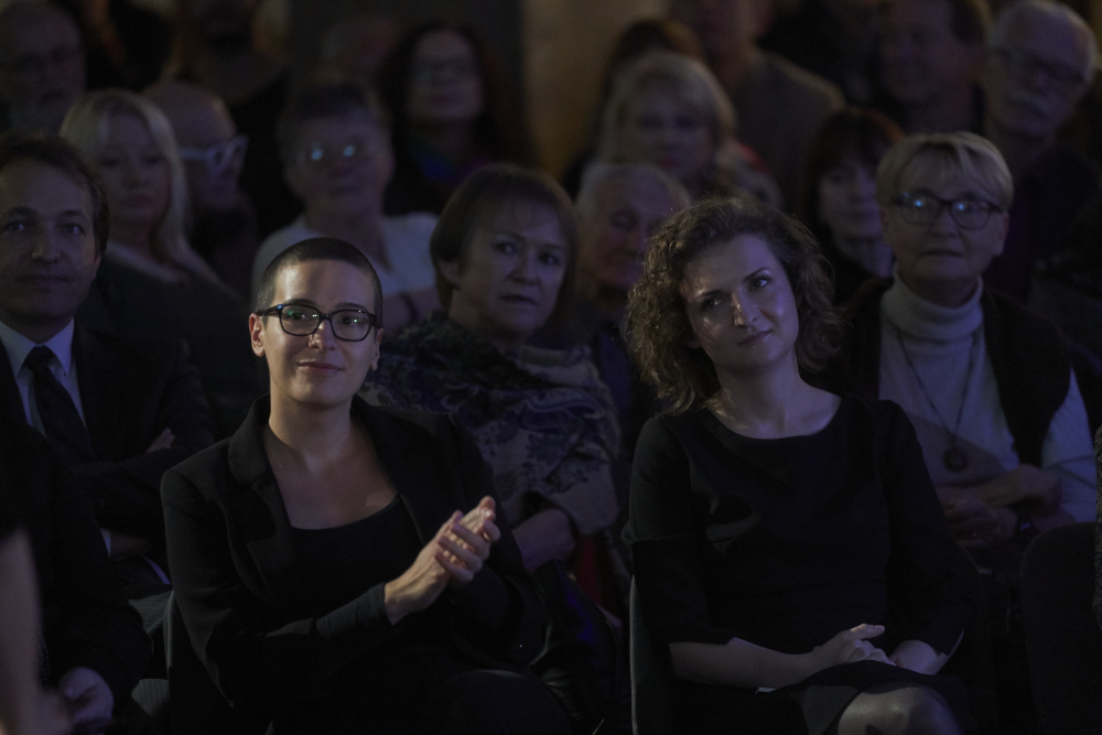 Kilka rzędów siedzących osób w przyciemnionej sali; na pierwszym planie po lewej osoba z bardzo krótkimi włosami, okularami i czarnym strojem klaszcze, obok niej osoba o kręconych włosach w czarnym ubraniu siedzi z rękami złożonymi na kolanach, w tle liczni widzowie, niektórzy w okularach i szalikach.