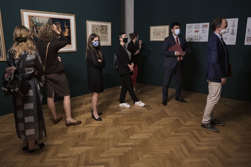 Grupa osób w maseczkach ustawiona w odstępach wewnątrz galerii sztuki, stojąca na jodełkowej drewnianej podłodze przed ciemnozieloną ścianą z oprawionymi rysunkami i plakatami.