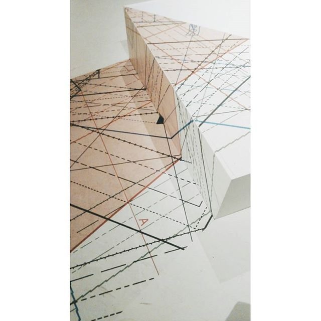 Zbliżenie na złożony biało‑beżowy papierowy model z nadrukowanymi czarnymi, przerywanymi i kolorowymi liniami tworzącymi złożoną siatkę geometrycznych, ukośnych wzorów i ostrych krawędzi.