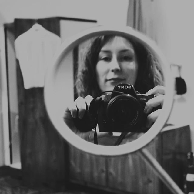 Czarno-białe zdjęcie odbicia osoby trzymającej aparat Sony widoczne w małym okrągłym lustrze, w tle drewniana szafa z powieszoną białą koszulą oraz fragmenty mebli i zasłony.