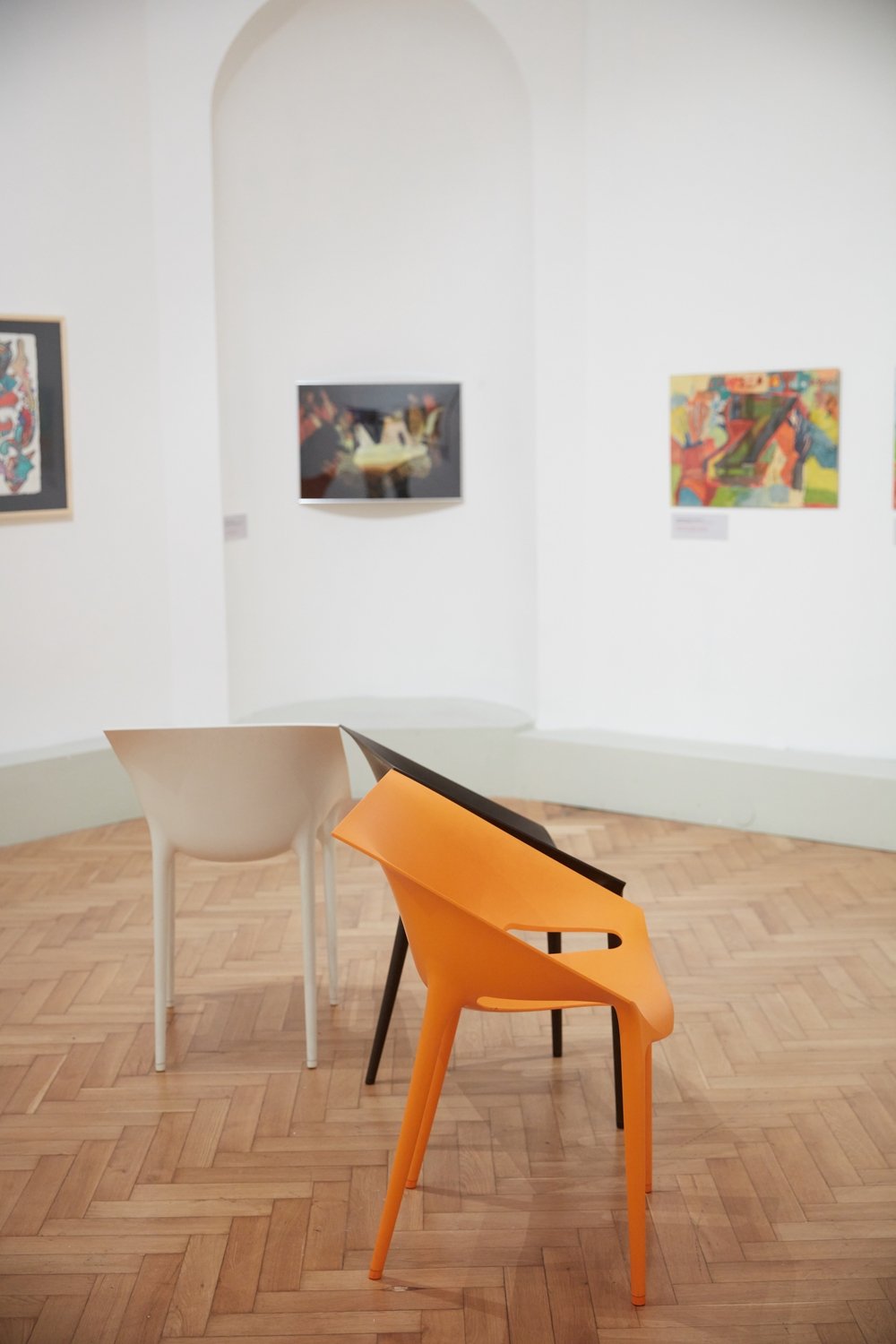 Trzy nowoczesne plastikowe krzesła — pomarańczowe z przodu, czarne w środku i białe z tyłu — zachodzą na siebie i stoją na jasnej drewnianej podłodze w galerii z białymi ścianami, łukowatą niszą i kilkoma obrazami na ścianach.