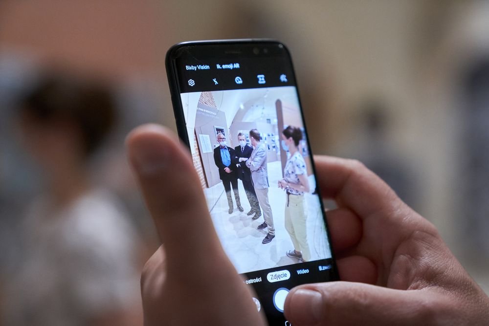 Zbliżenie na dłonie trzymające smartfon; na ekranie w trybie aparatu widać podgląd czterech osób stojących w jasnym, łukowym wnętrzu (jedna osoba w maseczce) oraz ikony aparatu i przycisk migawki z napisem "Zdjęcie".