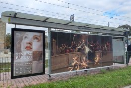Szklany przystanek tramwajowy z metalową wiatą i dwiema dużymi reklamami — po lewej plakat z twarzą kobiety i napisem "GENIUSZ BAROKU", po prawej duża grafika przedstawiająca scenę z wieloma postaciami; w tle tory tramwajowe, sieć trakcyjna, chodnik i pas trawy.