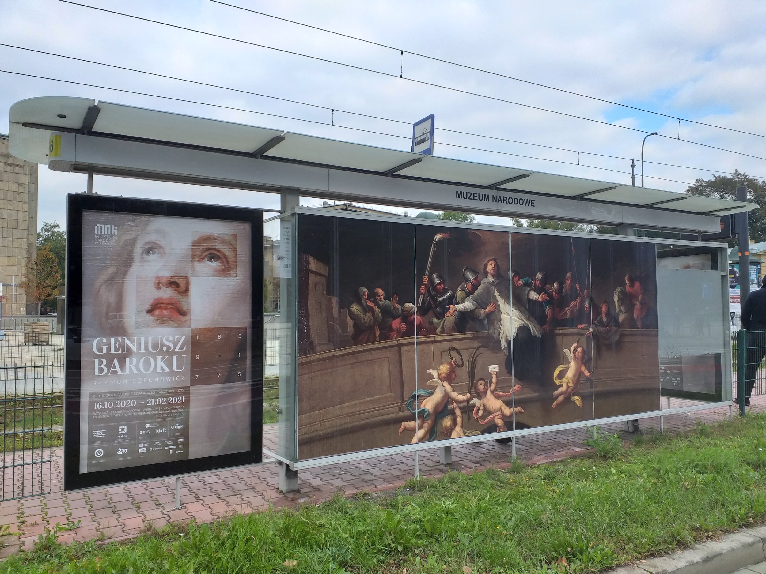 Przystanek tramwajowy z przeszkloną wiatą oznaczony napisem "Muzeum Narodowe", z dużym pionowym plakatem wystawy "Geniusz Baroku" po lewej pokazującym zbliżenie twarzy oraz szeroką wielopanelową reprodukcją barokowego obrazu z postaciami i puttami na środkowych i prawych szybach, na pierwszym planie chodnik i trawnik, w tle tory tramwajowe i zachmurzone niebo.