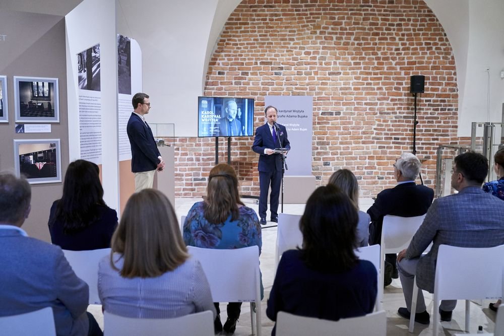 Mężczyzna w garniturze przemawia przy mikrofonie na tle ceglanej ściany i monitora z dużym zdjęciem, obok stoi drugi mężczyzna przy panelach wystawy, a kilkanaście osób siedzi na białych krzesłach, oglądając prezentację w jasnej sali muzealnej.
