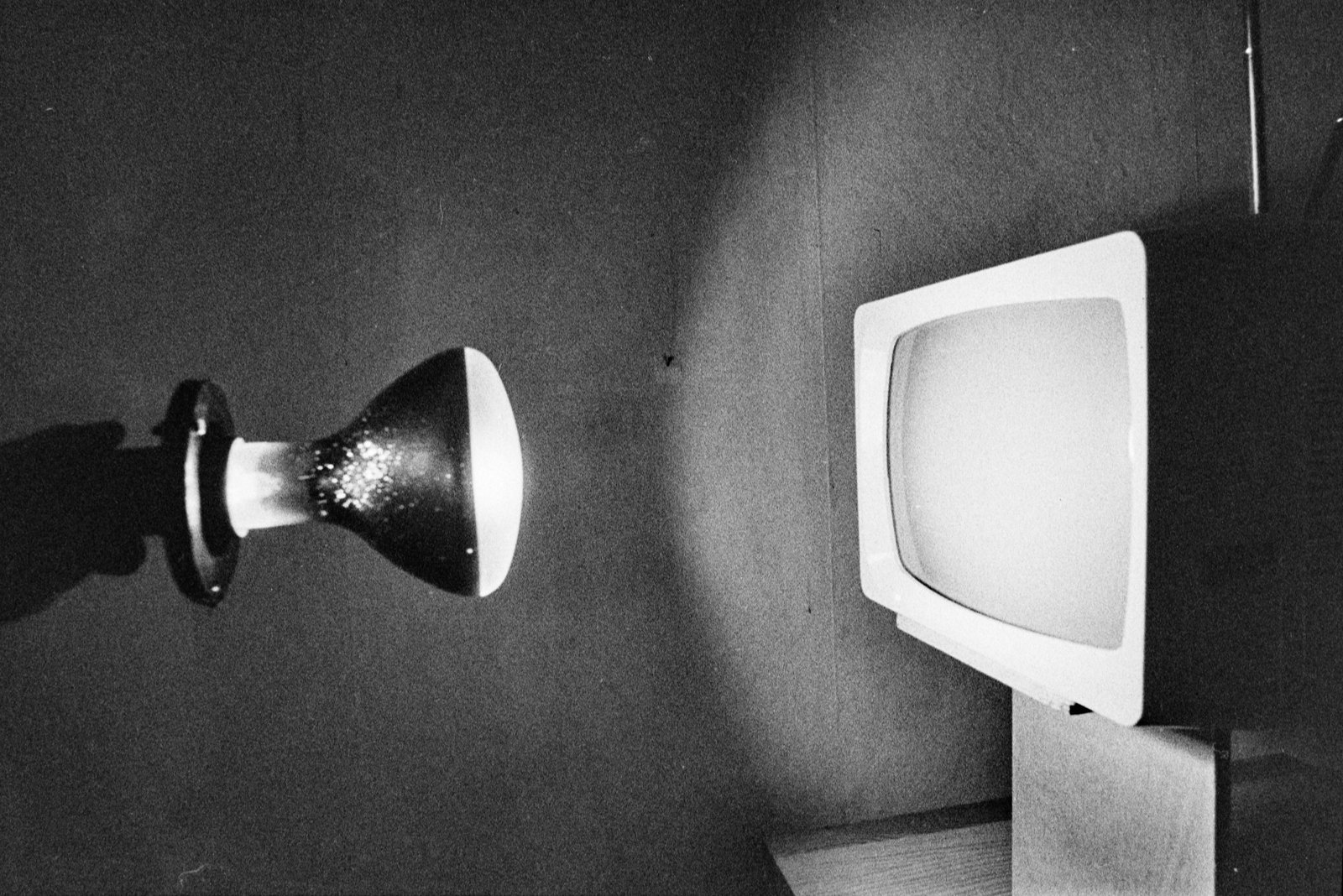 Czarno-białe, ziarniste zdjęcie przedstawiające po lewej przymocowaną do ściany lampę z dużą żarówką, po prawej telewizor kineskopowy na podstawie i między nimi widoczną plamę światła padającą na ścianę.