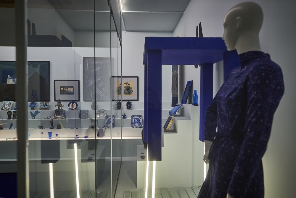 Witryna wystawowa z przeszkloną gablotą zawierającą na półkach drobne niebiesko-białe naczynia i dekoracje, kilka oprawionych obrazów na tylnej ścianie, nad nimi odwrócona niebieska konstrukcja przypominająca stół oraz po prawej stronie manekin w ciemnoniebieskiej sukience; u dołu widoczne pionowe pasy świetlne i odbicia na szkle.