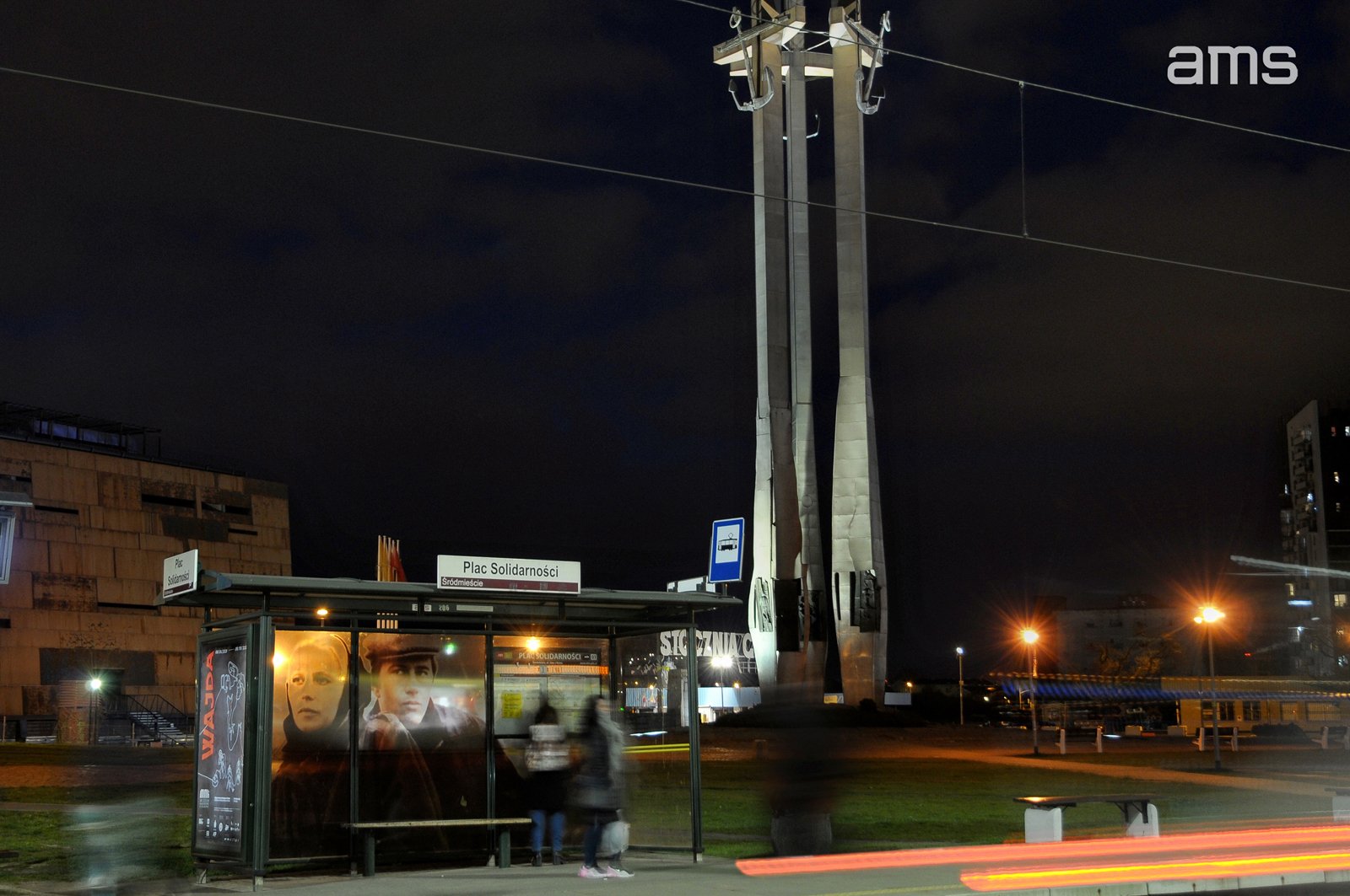 Nocne zdjęcie miejskiego przystanku z wiatą i podświetloną reklamą z dwiema twarzami, znakiem "Plac Solidarności", rozmytymi sylwetkami osób, śladami świateł przejeżdżających pojazdów i oświetlonym wysokim pomnikiem z trzema filarami na tle ciemnego nieba; w prawym górnym rogu logo "ams".
