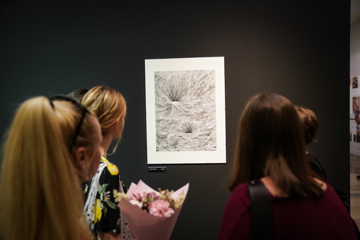 Cztery osoby widziane od tyłu oglądają oprawiony czarno-biały rysunek o gęstych, skończonych liniach zawieszony na ciemnej ścianie galerii; jedna z osób trzyma różowy bukiet kwiatów.