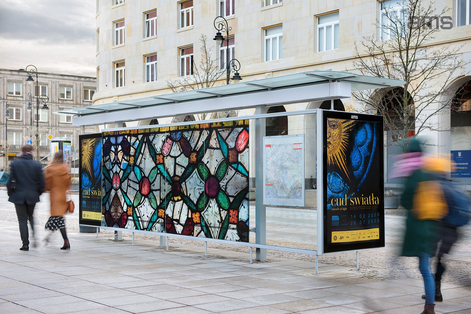 Przystanek autobusowy ze szklanym dachem na miejskim placu, z długą kolorową grafiką przypominającą fragment witraża oraz bocznym podświetlanym plakatem z napisem "cud światła", rozmazowane sylwetki osób przechodzących obok i w tle kamieniczne budynki, latarnie oraz nagie drzewa.