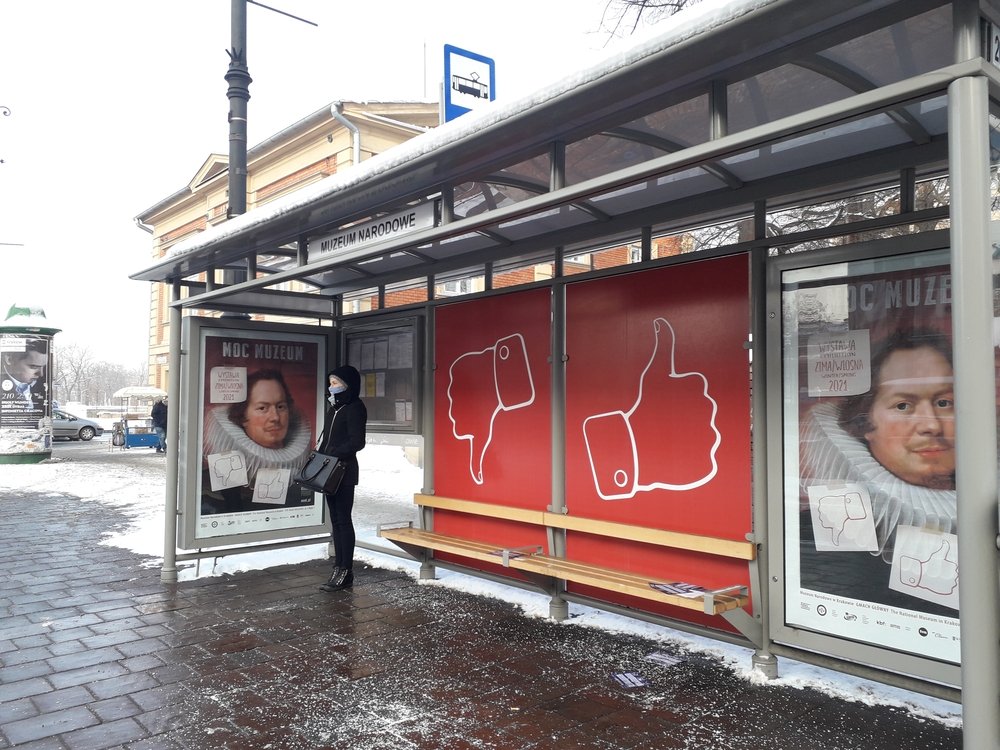 Przystanek autobusowy z zadaszeniem i widocznym napisem Muzeum Narodowe, czerwonymi panelami z dużymi ikonami kciuka w górę i w dół, drewnianą ławką oraz plakatami z portretem mężczyzny w renesansowym kołnierzu; przy przystanku stoi osoba w czarnym płaszczu i maseczce, na ziemi i dachu śnieg, w tle budynek i zaśnieżony chodnik.