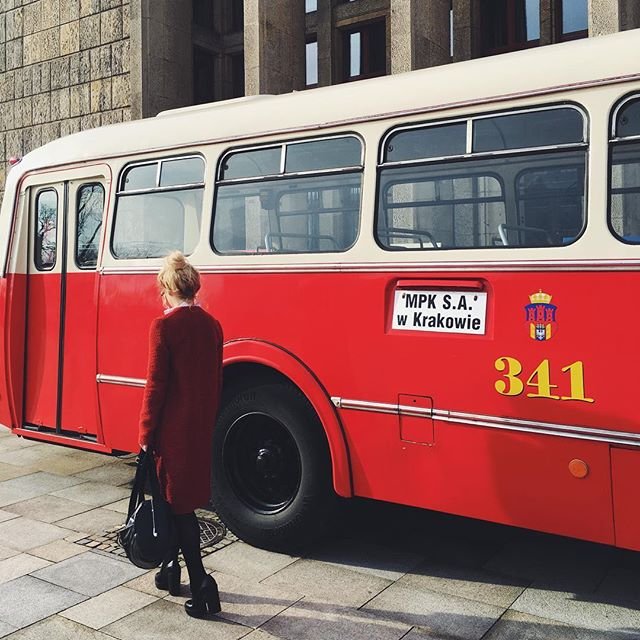 Czerwony autobus z białym dachem oznaczony numerem 341 i napisem "MPK S.A. w Krakowie" stoi przed kamienną fasadą, obok niego stoi odwrócona kobieta w czerwonym płaszczu trzymająca czarną torbę.