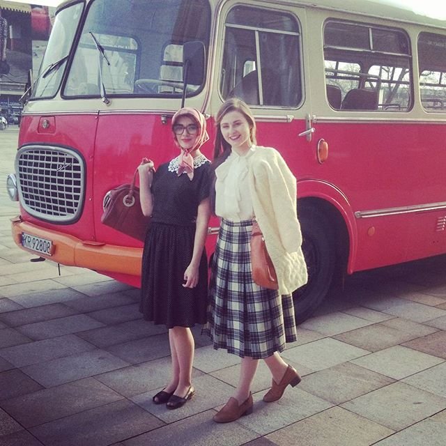 Dwie kobiety w retro ubraniach stoją przed czerwono‑kremowym zabytkowym autobusem na brukowanym placu; jedna w ciemnej sukience, apaszce na głowie i okularach trzyma brązową torebkę, druga ma jasną bluzkę, kremowy sweter, kratowaną spódnicę i brązową torebkę.