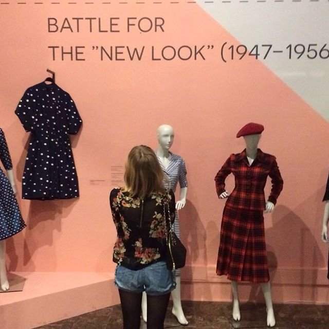 Osoba z tyłu, w kwiatowej bluzce, krótkich jeansowych spodenkach i czarnych rajstopach, stoi przed różową ścianą wystawy z dużym napisem BATTLE FOR THE "NEW LOOK" (1947–1956), oglądając manekiny prezentujące sukienki — po lewej granatową z białymi grochami powieszoną na wieszaku, na środku biało‑niebieską w paski, a po prawej czerwoną w kratę z beretem.