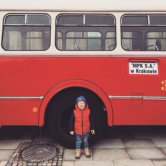 Małe dziecko w czerwonej pikowanej kamizelce, niebieskiej czapce, dżinsach i żółtych butach stoi przed dużym czerwonym autobusem pod nadkolem koła, na bocznej ścianie autobusu widoczny napis MPK S.A. w Krakowie, a na chodniku przed nim właz kanalizacyjny.