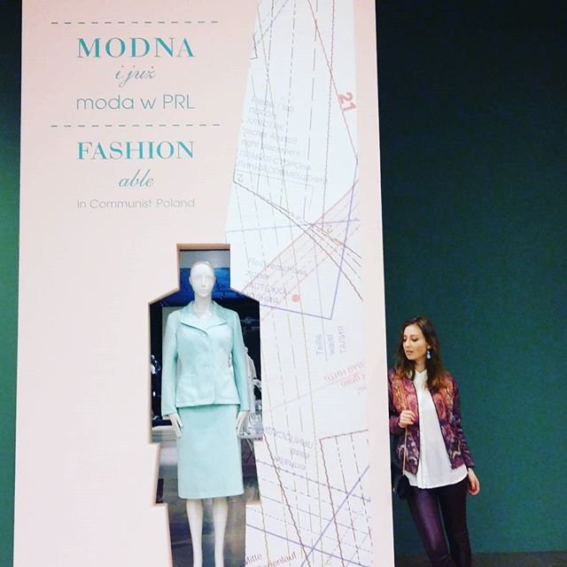 Różowy pionowy panel wystawy z rysunkami wykrojów i widocznymi napisami "MODNA i już moda w PRL" oraz "FASHION able in Communist Poland", w wycięciu którego stoi manekin ubrany w miętowy żakiet i spódnicę, a po prawej stoi kobieta w ciemnofioletowej kurtce i białej bluzce na tle ciemnozielonej ściany.