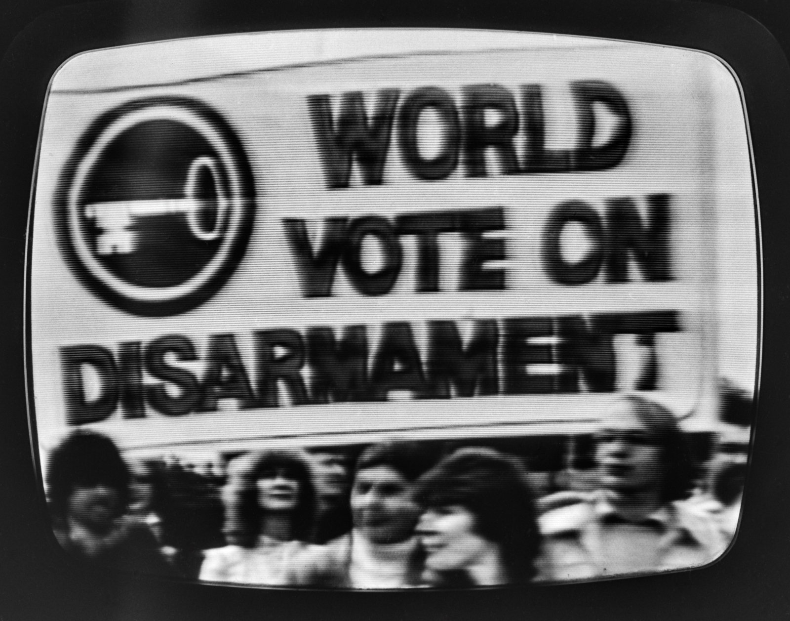 Czarnobiały kadr ekranu telewizora pokazujący duży transparent z napisem WORLD VOTE ON DISARMAMENT i symbolem klucza w okręgu po lewej, poniżej rozmyty tłum osób oraz widoczne poziome linie i zaokrąglone narożniki ekranu.
