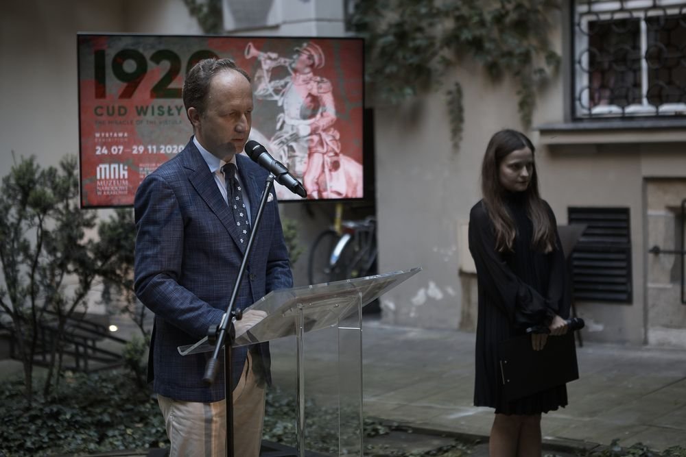 Mężczyzna w niebieskiej kratowanej marynarce stoi przy przezroczystym pulpicie i mikrofonie na zewnętrznym dziedzińcu; za nim duży czerwony plakat z napisem 1920 CUD WISŁY i datami wystawy, a po prawej stoi kobieta w czarnej sukience trzymająca teczkę i mikrofon, w tle ściana budynku, okno z kratą i roślinność.