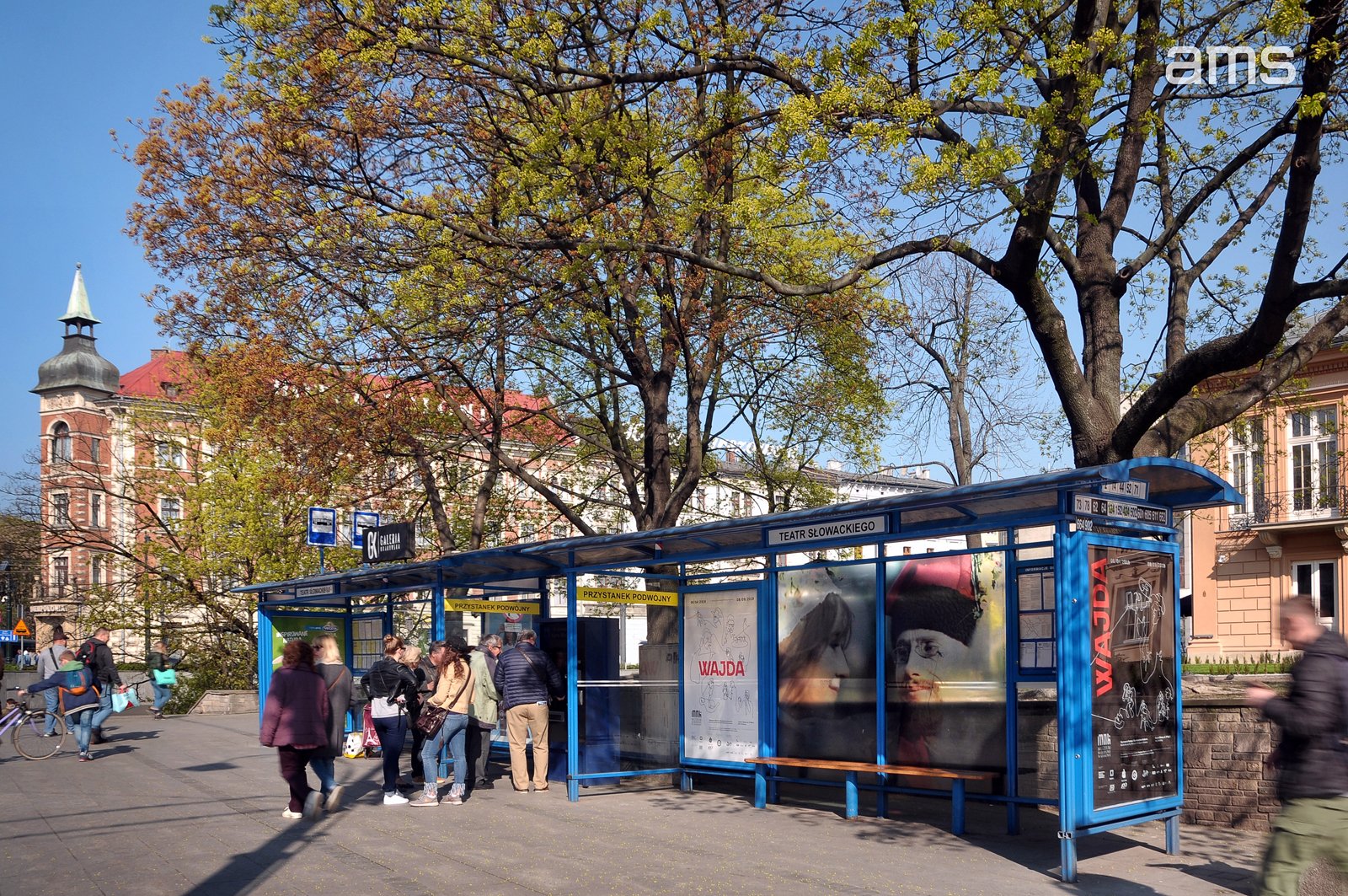 Przestrzenny miejski przystanek z niebieską wiatą oznaczony "TEATR SŁOWACKIEGO", z kilkoma osobami czekającymi przy szklanych panelach z plakatami (m.in. "WAJDA"), drewnianą ławką, dużymi drzewami z młodymi zielono‑żółtymi liśćmi, zabytkowymi kamienicami z czerwoną wieżą w tle, rowerem i przechodniami na chodniku pod jasnym niebieskim niebem; w prawym górnym rogu widoczne logo "ams".