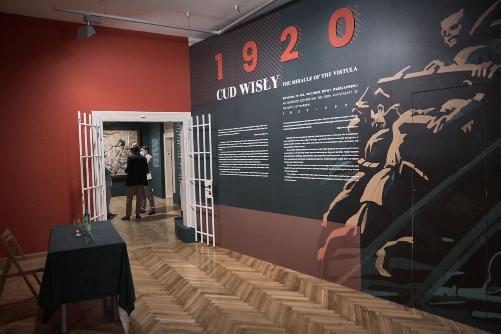 Wnętrze sali wystawowej z dużą ciemną ścianą z napisem "1920 CUD WISŁY" i dużą grafiką postaci w mundurze po prawej, białą metalową kratą wokół przejścia, dwiema osobami rozmawiającymi w drzwiach, stolikiem z zielonym obrusem i butelkami po lewej oraz jasną parkietową podłogą.