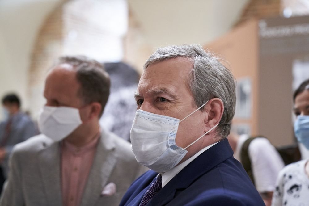 Mężczyzna w siwych włosach ujęty z profilu w pierwszym planie, w chirurgicznej masce i granatowym garniturze, a w tle rozmyte postacie również w maseczkach i tablice ekspozycyjne.